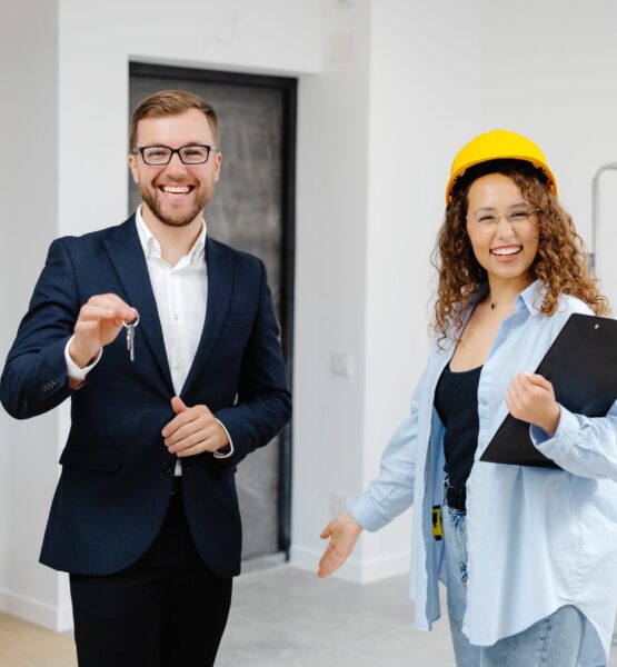 Female designer in helmet with documents giving keys from apartment to young businessman while standing in room.